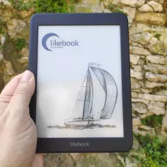 Une main tient un e-reader Likebook affichant un voilier. L'appareil est devant un mur de pierres couvert de végétation.