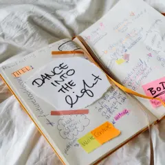 Un carnet ouvert avec des notes manuscrites sur la création d'un livre, comme "DANCE INTO THE LIGHT". On y voit des idées de titres, de marketing, et la question "a quoi sert un pseudo" est implicite dans la planification.