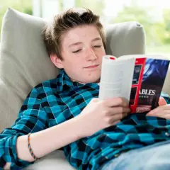 Un adolescent aux taches de rousseur lit un livre captivant, plongé dans un monde d'aventures. Il est confortablement installé sur un canapé, entouré de livres pour adolescents.