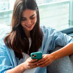 Une jeune femme souriante lit un livre à lire sur téléphone, confortablement installée sur un canapé gris.