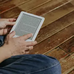 Une personne lit sur une tablette encre électronique, assise sur un plancher en bois.