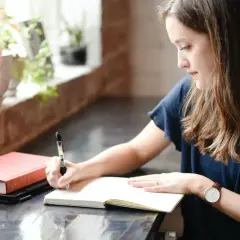 Une jeune femme écrit dans un carnet, peut-être le début de sa propre biographie. À côté, un livre rouge et une tasse de café.