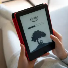 Une personne lit sur une liseuse Kindle, affichant une barre de progression et une silhouette sous un arbre. Un guide d'utilisation Kindle est à portée de main.