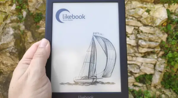 Une main tient un e-reader Likebook affichant un voilier. L'appareil est devant un mur de pierres couvert de végétation.