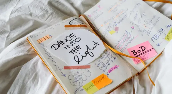 Un carnet ouvert avec des notes manuscrites sur la création d'un livre, comme "DANCE INTO THE LIGHT". On y voit des idées de titres, de marketing, et la question "a quoi sert un pseudo" est implicite dans la planification.