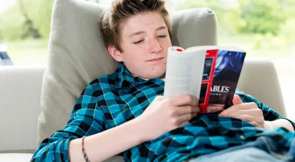 Un adolescent aux taches de rousseur lit un livre captivant, plongé dans un monde d'aventures. Il est confortablement installé sur un canapé, entouré de livres pour adolescents.