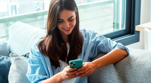 Une jeune femme souriante lit un livre à lire sur téléphone, confortablement installée sur un canapé gris.