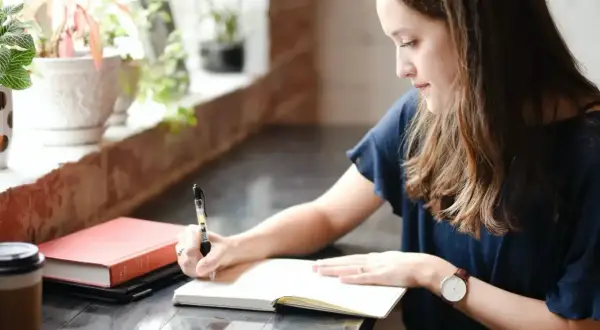 Une jeune femme écrit dans un carnet, peut-être le début de sa propre biographie. À côté, un livre rouge et une tasse de café.