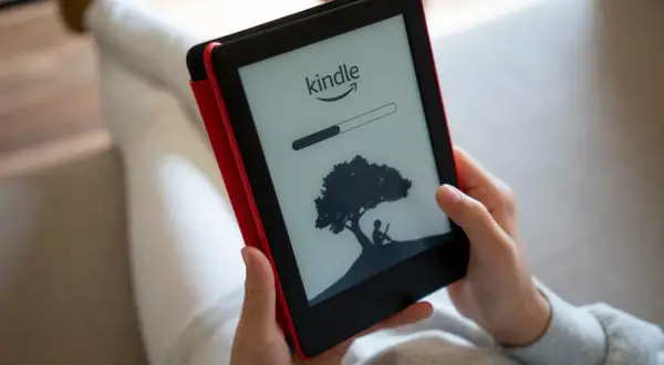 Une personne lit sur une liseuse Kindle, affichant une barre de progression et une silhouette sous un arbre. Un guide d'utilisation Kindle est à portée de main.