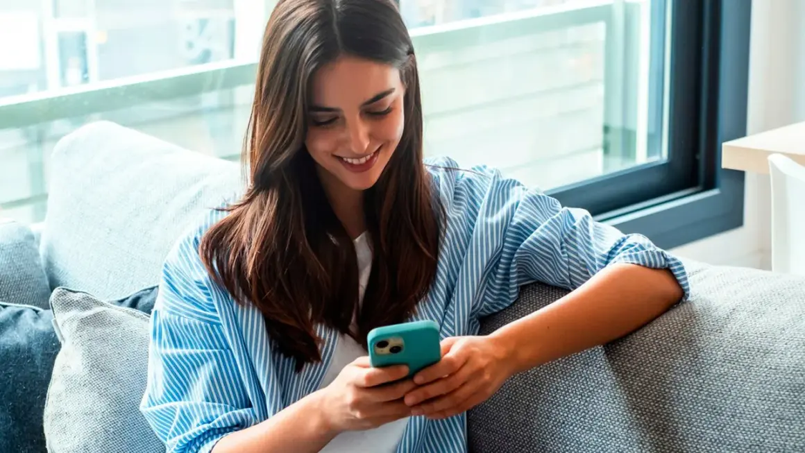 Une jeune femme souriante lit un livre à lire sur téléphone, confortablement installée sur un canapé gris.