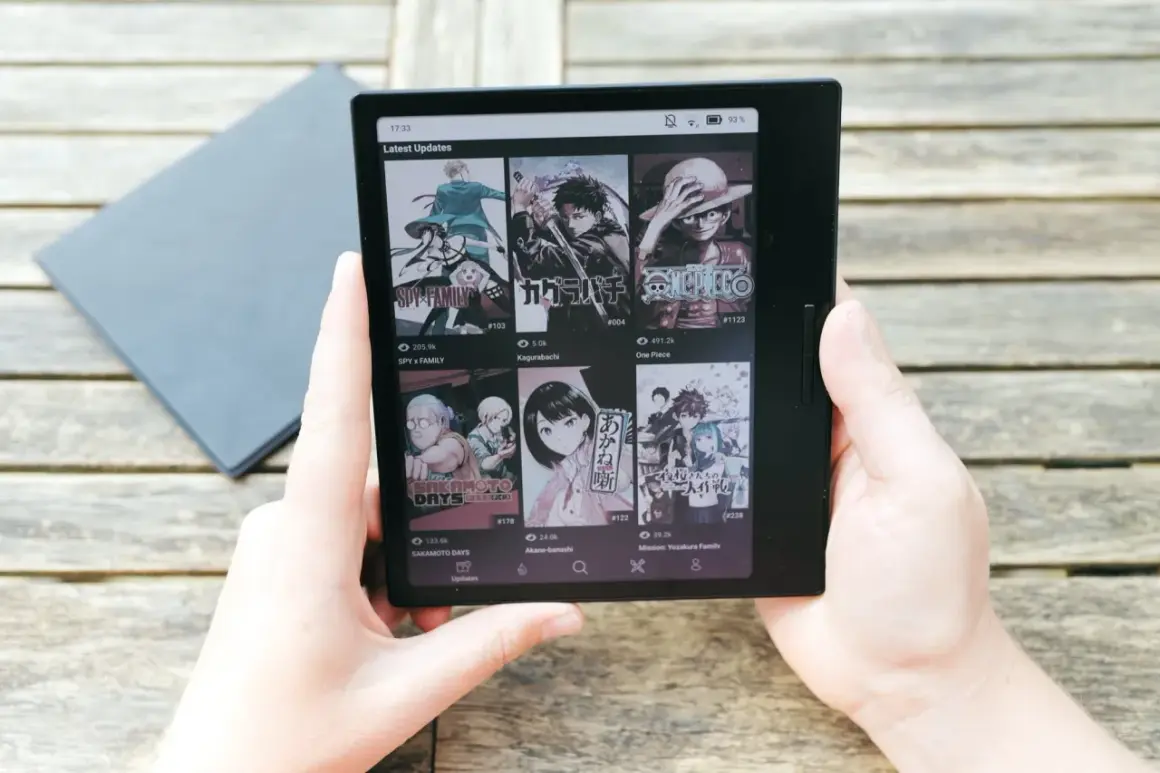 Mains tenant une liseuse affichant des mises à jour de mangas, dont Spy x Family, Kagurabachi et One Piece, sur un fond de table en bois. Le kaleido 3 offre une lecture claire.