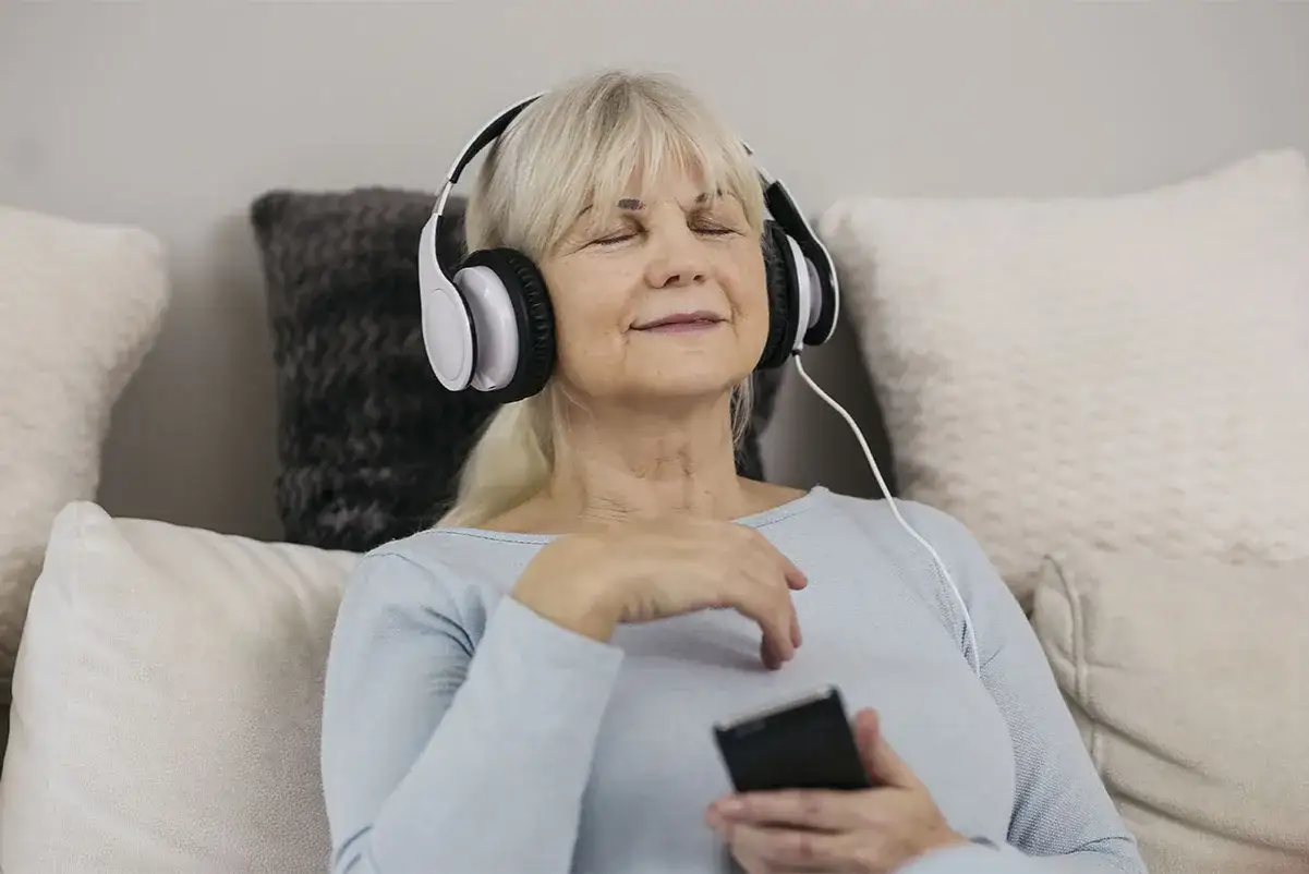 Une femme âgée écoute un livre audio gratuit sur son téléphone, les yeux fermés, détendue sur un canapé.