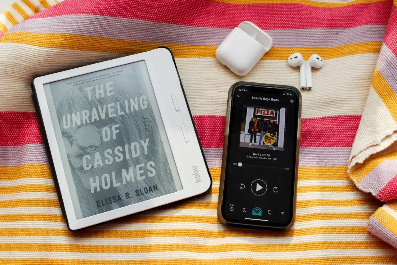 Un livre t&eacute;moignage sur un Kobo, un smartphone avec un podcast et des &eacute;couteurs sans fil, le tout sur une serviette ray&eacute;e.