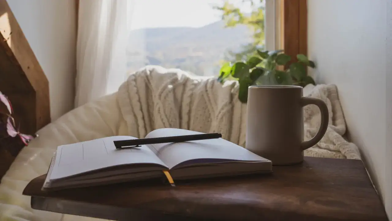 Un carnet ouvert avec un stylo, une tasse fumante et une couverture douillette pr&egrave;s d'une fen&ecirc;tre. Un moment parfait pour un feel good livre.