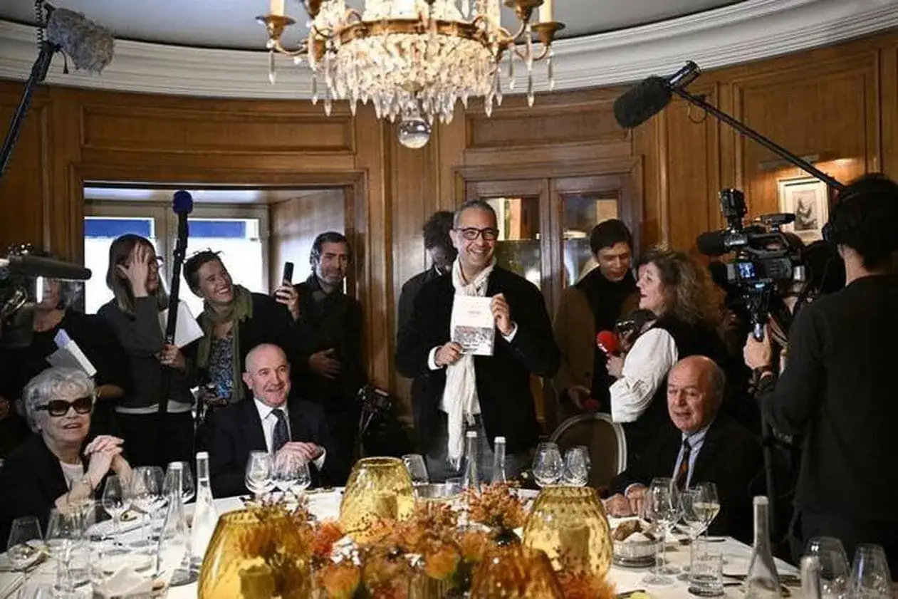 Un homme souriant pr&eacute;sente un livre, entour&eacute; de journalistes et de cam&eacute;ras, lors d'une annonce de prix litt&eacute;raire.