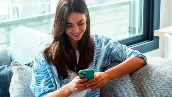 Une jeune femme souriante lit un livre à lire sur téléphone, confortablement installée sur un canapé gris.