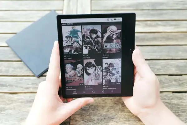 Mains tenant une liseuse affichant des mises à jour de mangas, dont Spy x Family, Kagurabachi et One Piece, sur un fond de table en bois. Le kaleido 3 offre une lecture claire.